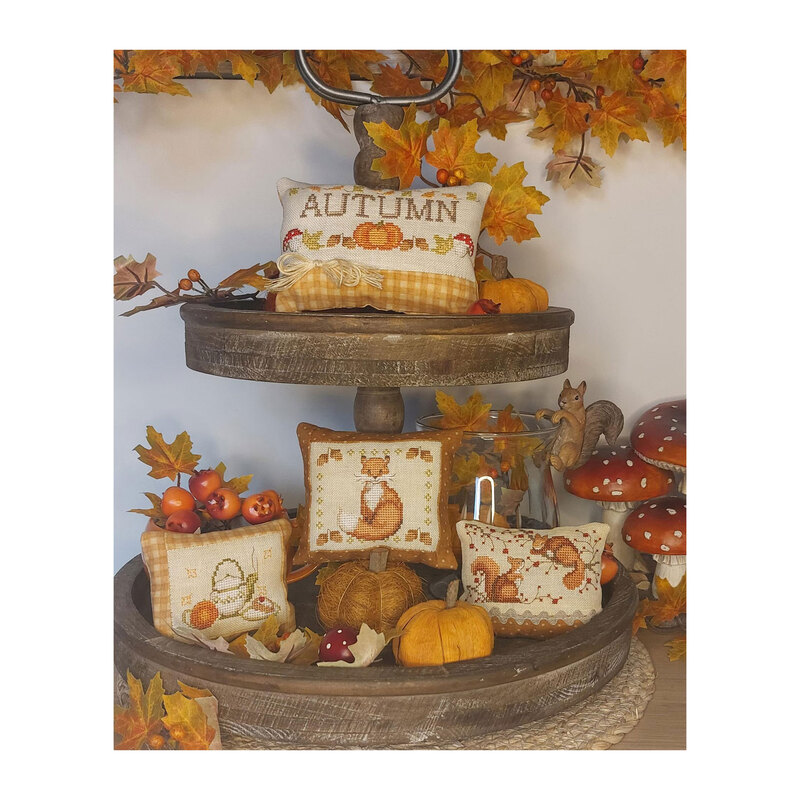 Photo of the four Autumn Pillow designs stacked on a tower of leaves, pumpkins, berries, and a squirrel hanging on a vase.