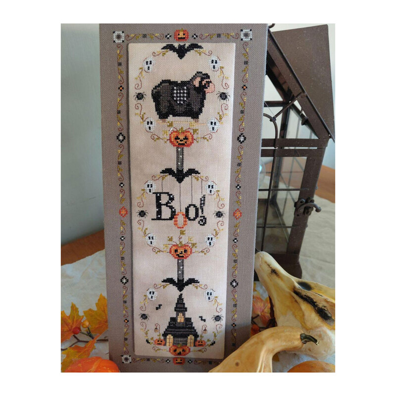 Enhanced photo of the finished Boo! cross stitch pattern leaning against a steel lantern next to yellow squash and maple leaves.