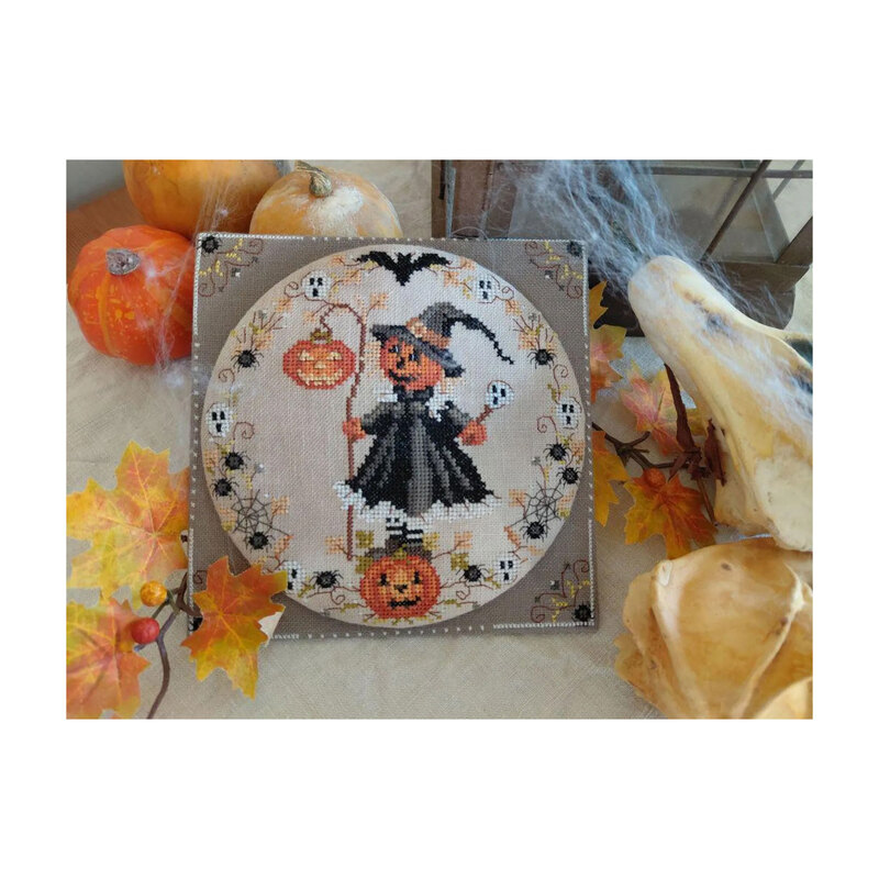 Photo of the finished Halloween - Pumpkin Witch cross stitch pattern finished in a circle hoop on a natural colored linen fabric board next to pumpkins and autumn leaves.