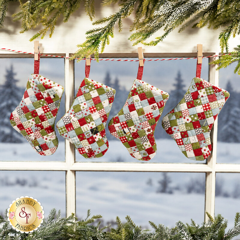 The four completed Fast Fusible Patchwork Stockings colored in wintry prints from the Chilly collection by Moda Fabrics, hung on a striped string by clothespins in a window that looks over a snowy scene; Christmas garlands peek into frame from the top and bottom.