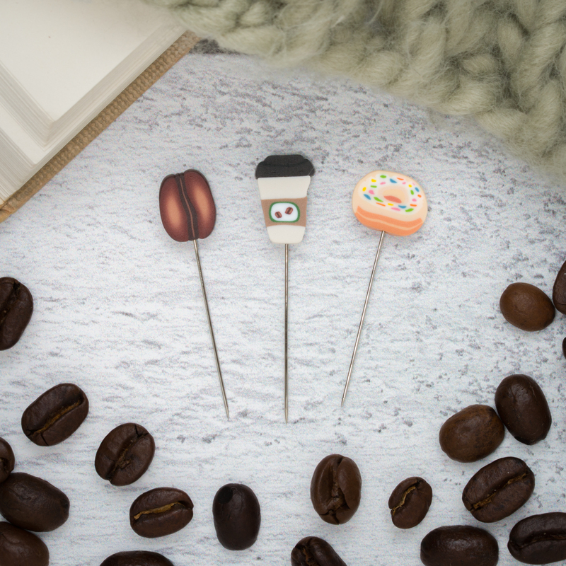 Three decorative straight pins topped with a coffee bean, takeaway coffee cup, and sprinkled donut.