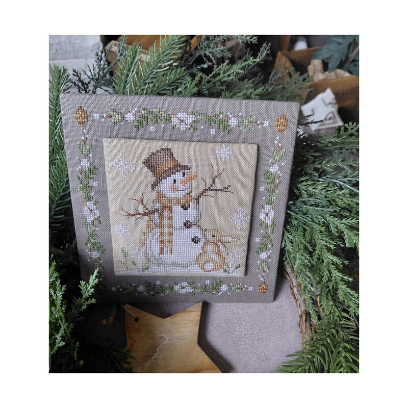 Photo of the cross stitch pattern with the finished pattern stitched onto a granite-gray finishing board in front of pine boughs.