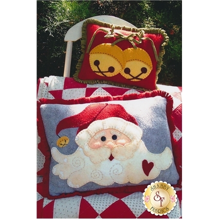 The front of the Jingle Bell Santa Pillows pattern featuring two holiday pillows, a cheery santa with a heart and a bell at the end of his cap, and two large holiday bells, on a red and white diamond quilt.