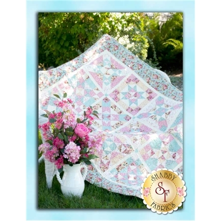 A colorful quilt with a star pattern is displayed in a garden setting alongside a white vase filled with pink flowers. The background features greenery.