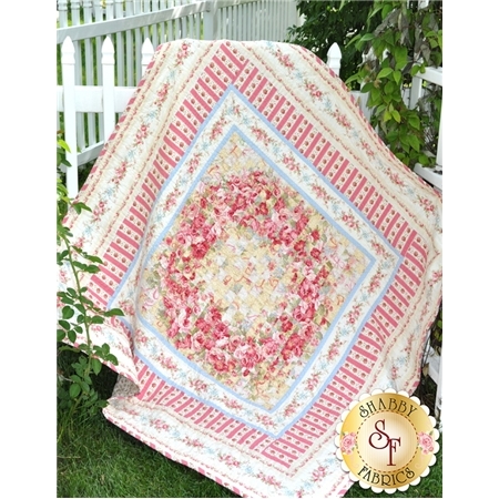 A colorful quilt with a floral pattern is draped over a fence in a grassy area. The quilt features a central diamond design surrounded by stripes and smaller floral motifs. The image includes a logo in the bottom right corner that reads Shabby Fabrics.