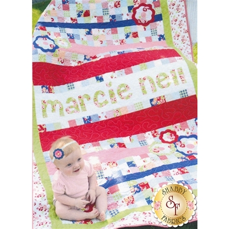 A colorful quilt featuring various patterns, including floral and geometric designs, with the name marcie nel stitched on it. A baby wearing a light pink shirt and a flower headband is sitting in front of the quilt. The bottom corner includes a logo for Shabby Fabrics.