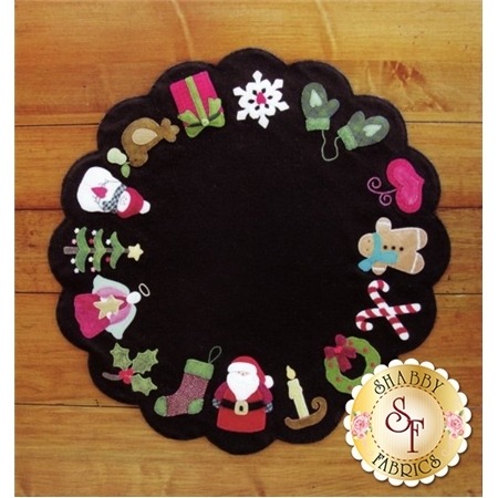 A circular black wool holiday mat featuring festive decorations along the edges, including Santa, a snowman, gingerbread cookies, candy canes, Christmas gifts, and holly. The design has a scalloped edge.