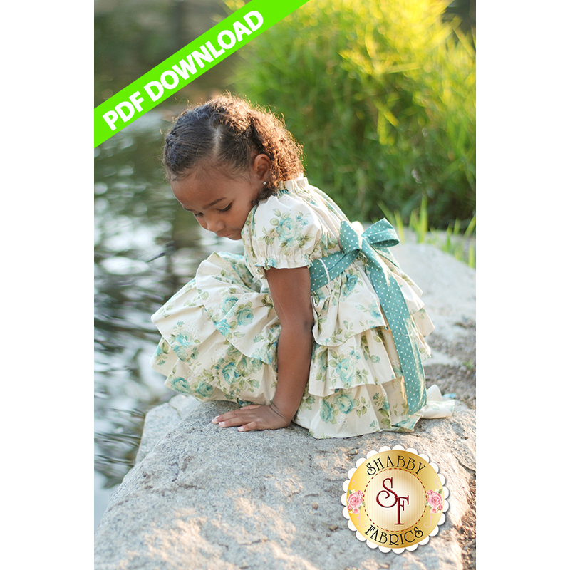 A young girl in a floral dress with a blue ribbon sits on a rock by a body of water, looking down. The background features greenery, and a PDF DOWNLOAD banner is in the top corner. The Shabby Fabrics logo is at the bottom center.