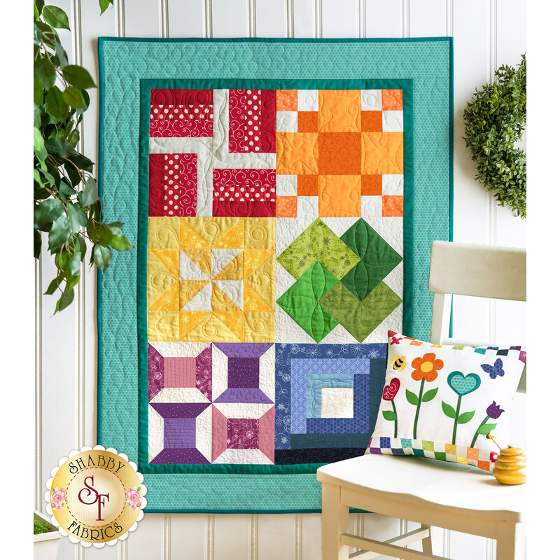 An image of a Learn To Quilt Beginner Quilt hanging on a wall surrounded by greenery.