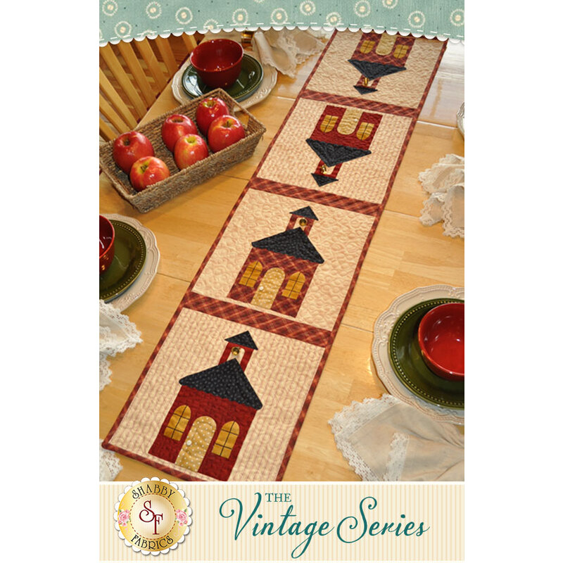 A table runner featuring four schoolhouse motifs, with apples and plates arranged on a wooden table.