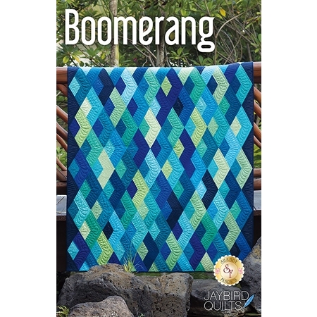 A colorful quilt titled Boomerang is displayed, featuring a diamond pattern with varying shades of blue and green. The quilt is hanging outdoors against a natural backdrop, and the logo for Jaybird Quilts is visible in the bottom right corner.