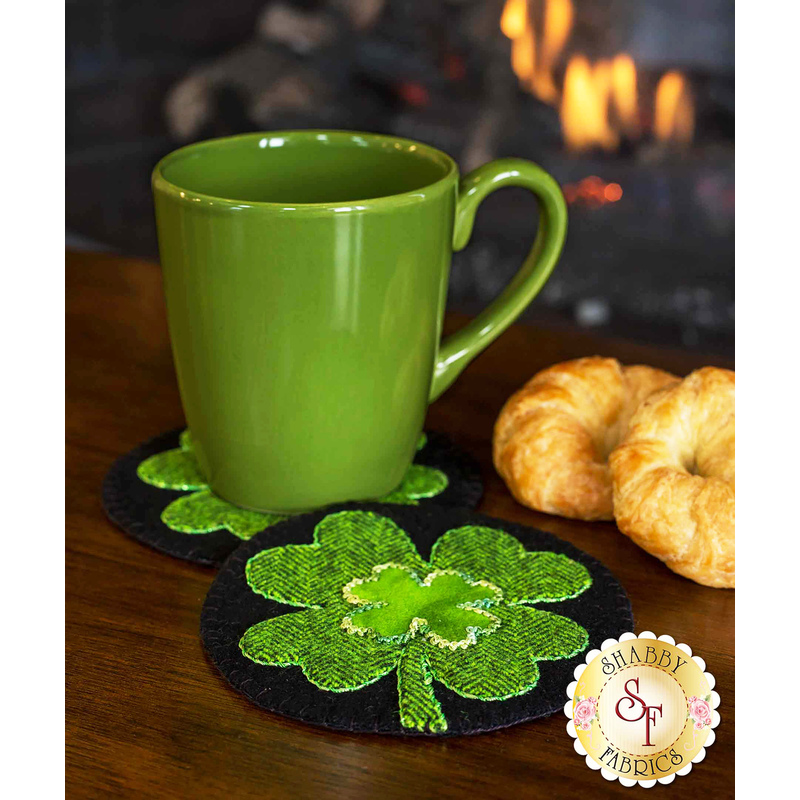 A green mug sits on a black circular coaster adorned with a bright green shamrock. Next to the mug is a flaky croissant. In the background, a fireplace is softly flickering.