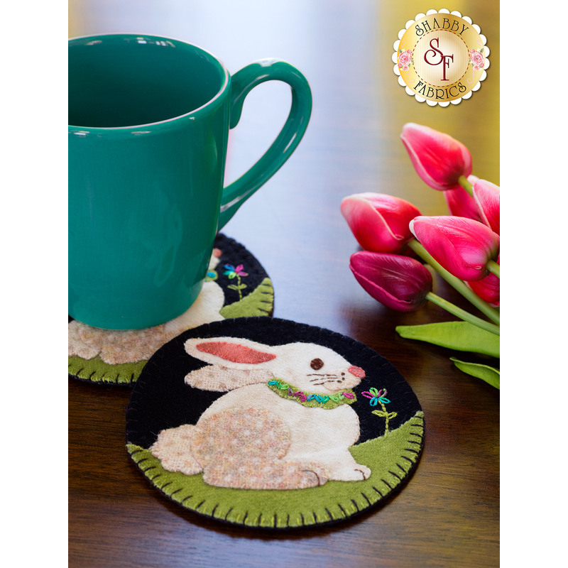 A green mug is placed on a decorative coaster featuring an illustration of a sitting bunny with a floral collar. Pink tulips and green leaves are arranged nearby on a wooden surface.