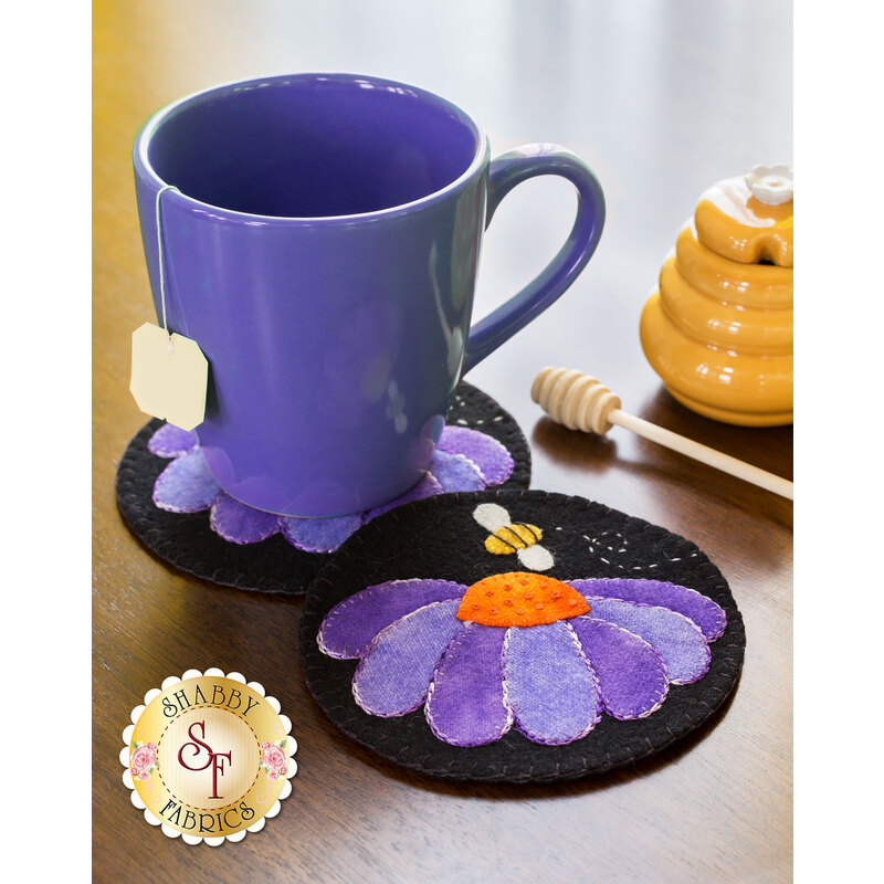 A purple mug sits on a decorative black coaster with a flower design. The coaster features petals in shades of purple and an orange center, with a small bee. Nearby, there is a honey jar and a honey dipper.