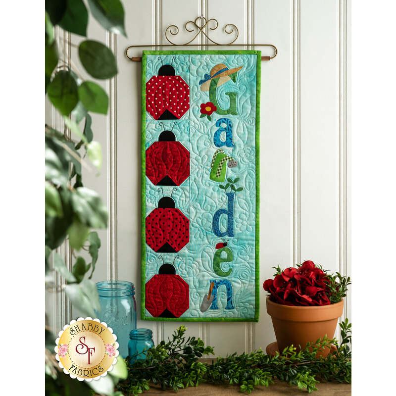 A colorful wall hanging with the word Garden in blue and green letters, surrounded by various ladybug designs. The background is light blue, and the hanging is decorated with a small hat and a flower. Below, there are potted flowers and green foliage.