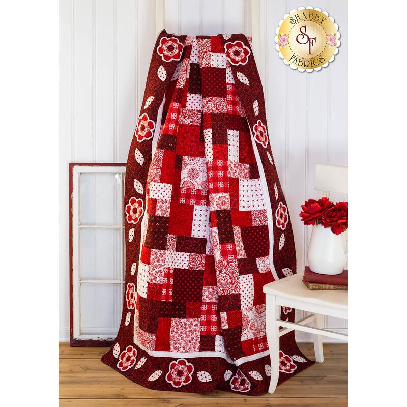 A colorful quilt in shades of red and white, featuring a patchwork design with floral accents, draped over a ladder in a bright room with wooden flooring.