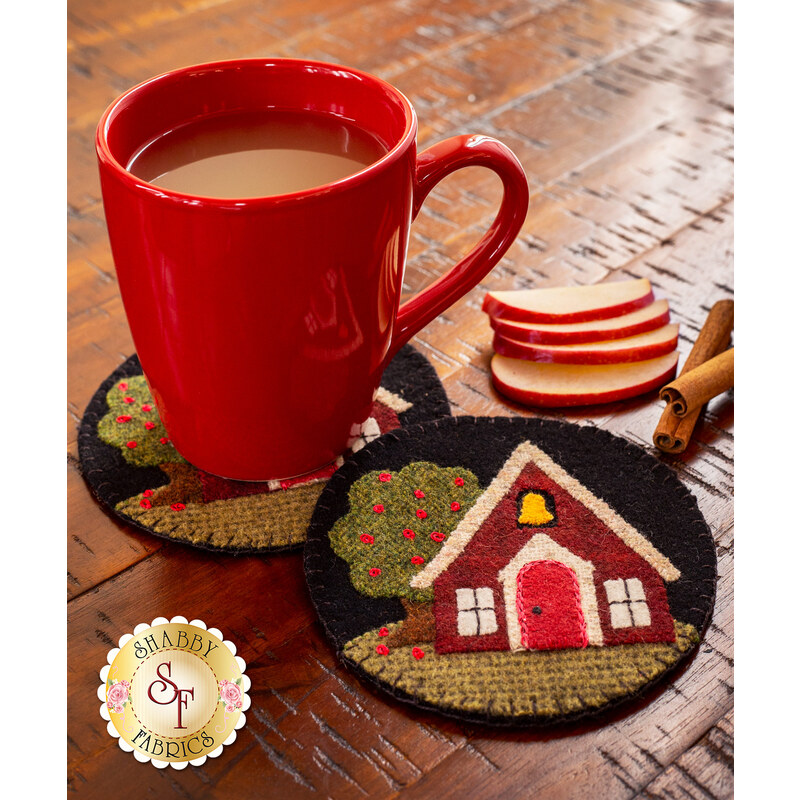 A red mug filled with coffee sits on a round black coaster featuring an illustrated house and trees. Next to the mug, there are several slices of apple and a cinnamon stick on a wooden table.