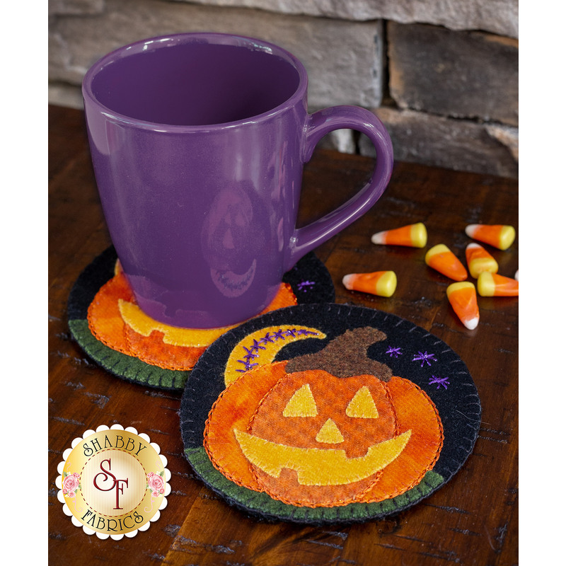 A purple mug is placed on two Halloween-themed coasters featuring pumpkins, surrounded by candy corn on a wooden surface.