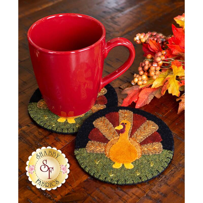 A red mug sits on two round coasters featuring a turkey design, surrounded by autumn leaves and berries on a wooden table.