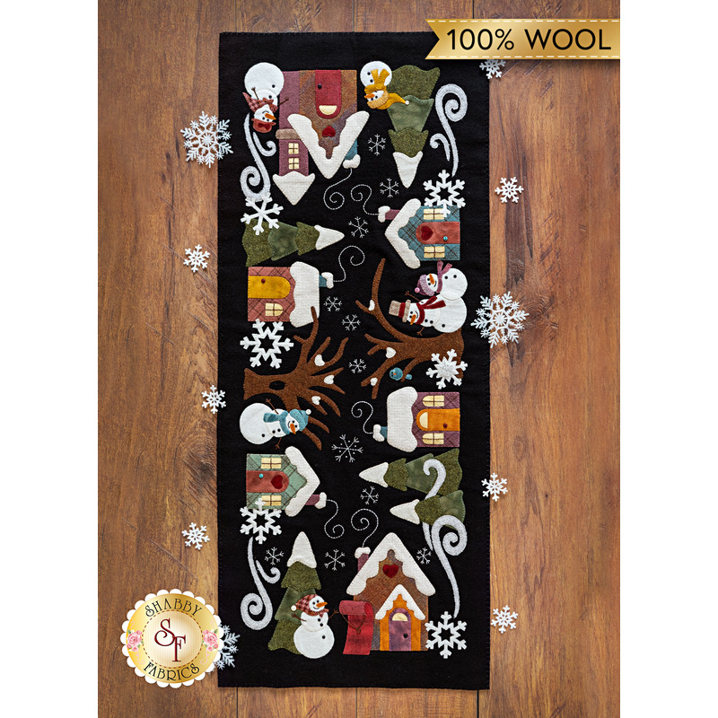 The completed Snowman Lane Flannel Table Runner, colored in vibrant Woolies Flannels, staged on a dark wooden table. A golden banner at the top reads 100% Wool. 