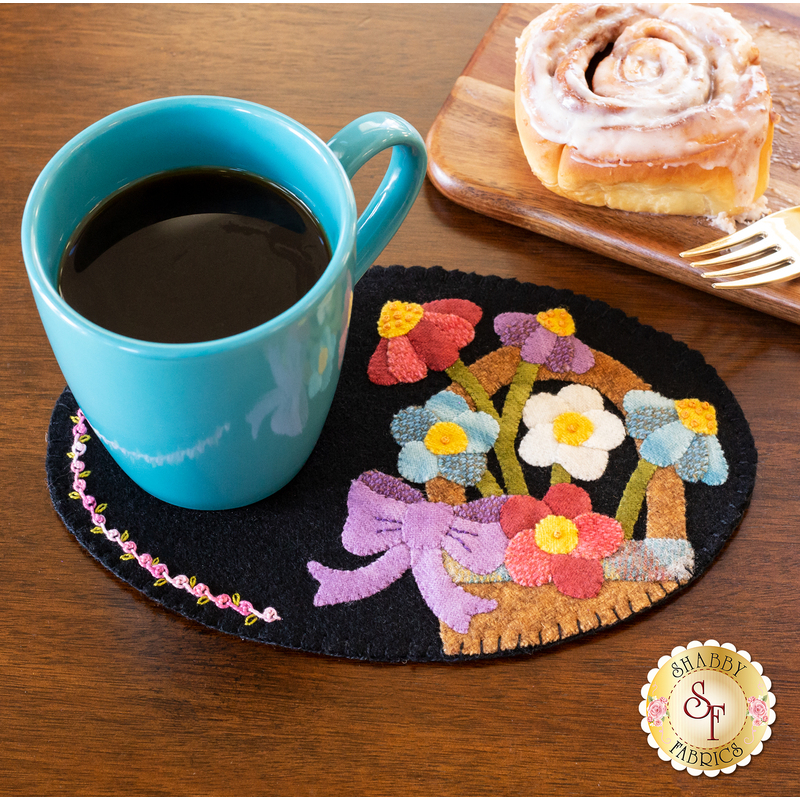 A turquoise coffee mug sits on a decorative black wool coaster featuring a colorful floral design, next to a wooden plate holding a cinnamon roll and a fork.