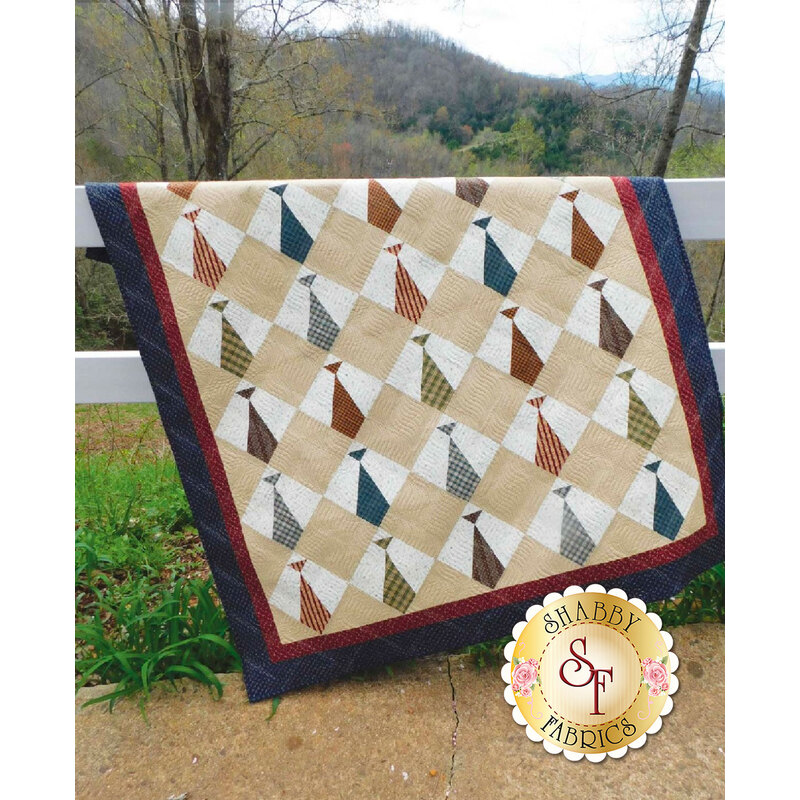 A patchwork quilt with a diamond pattern featuring colorful ties hangs on a fence in a natural landscape.