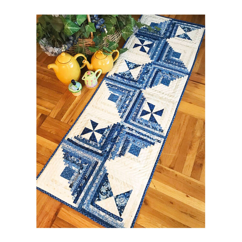 A blue and white quilted table runner laid on a wooden floor, with yellow teapots and a green teacup.